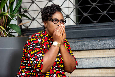 beautiful mature woman wearing eyeglasses with a traditional loincloth sitting sideways on the stairs and covering her mouth with her hands shocked by shameの写真素材