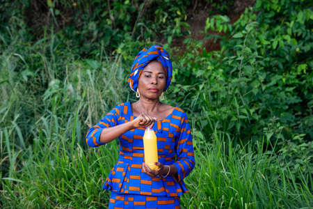 a beautiful mature african woman in traditional dress standing in the countryside holding a bottle of fruit juiceの写真素材
