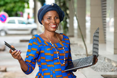 beautiful mature businesswoman in traditional outfit standing outdoors holding laptop and mobile phone looking at camera smiling.の写真素材