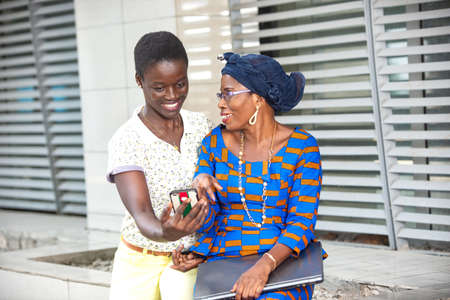 two beautiful adult businesswomen sitting with a mobile phone outdoors smiling with laptop in hand.の写真素材