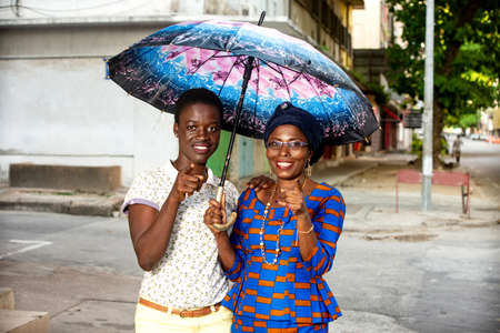 two beautiful adult women standing under an umbrella in the street and pointing fingers at something while smiling.の写真素材