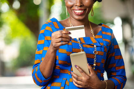 beautiful african businesswoman in traditional outfit standing outdoors holding mobile phone and credit card smiling.の写真素材