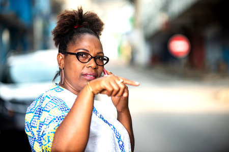 Beautiful mature woman in glasses standing outdoors communicating on cellphone and making finger...