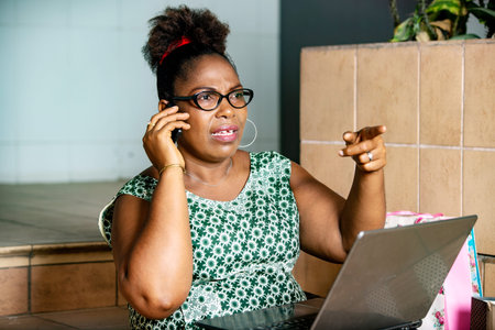 mature business woman sitting and talking on mobile phone with laptop on her legs and pointing...