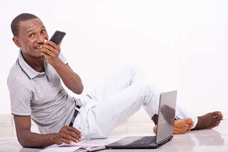a handsome student in a t-shirt sitting on the floor isolated on a white background talking on the cellphone and looking at the camera smiling.の写真素材