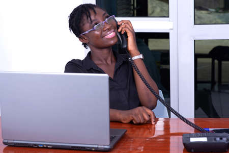 young businesswoman wearing glasses, sitting at the desk and talking on the mobile phone with eyes raised up and smiling.の写真素材