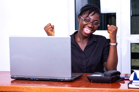beautiful young businesswoman sitting at the desk using a laptop and making a winning gesture with...