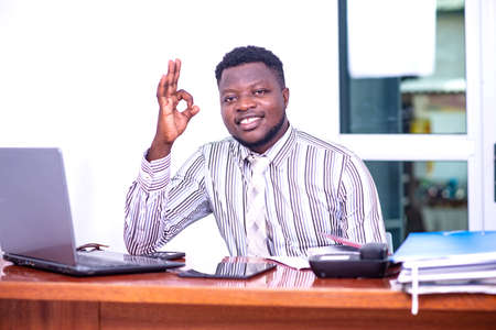 a young businessman in striped shirt sitting at the desk with laptop and showing okay sign while smilingの写真素材