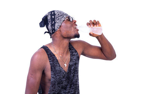 handsome thirsty young man wearing tank top and bandana over his head, drinking a bottle of mineral...