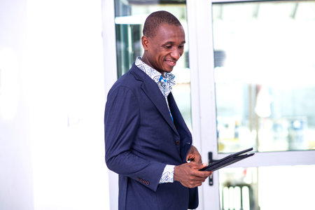 handsome young businessman standing at desk, wearing elegant suit, using digital tablet while...