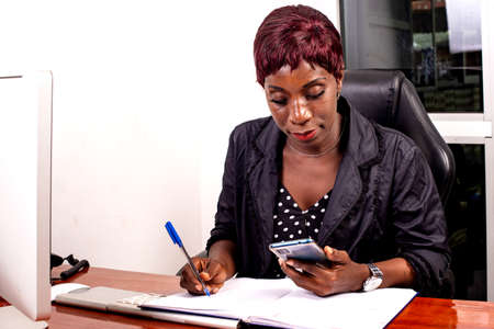 portrait of a beautiful adult businesswoman sitting at the desk and writing in the notebook using a mobile phone.の写真素材