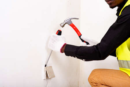 young male electrician wearing white glove and using hammer and screwdriver to install electrical outlets and switch.の写真素材