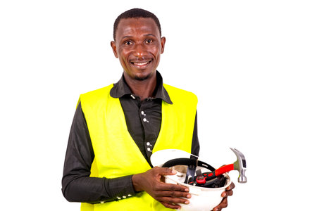 young man technician wearing green vest showing safety helmet with tools indoors while smiling.