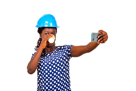 young female engineer wearing blue safety helmet and drinking paper cup of coffee while taking...