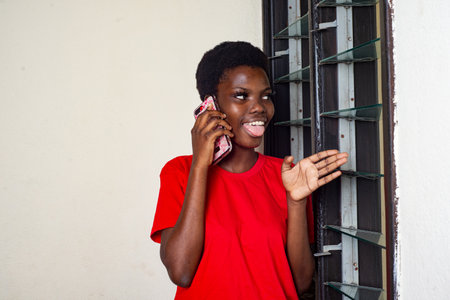 young beautiful girl in red t-shirt standing at home talking on the cellphone near the window sticking out her tongue.の写真素材