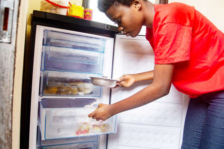 young beautiful girl in a red t-shirt standing in the kitchen looking for something to cook in the refrigerator.の写真素材