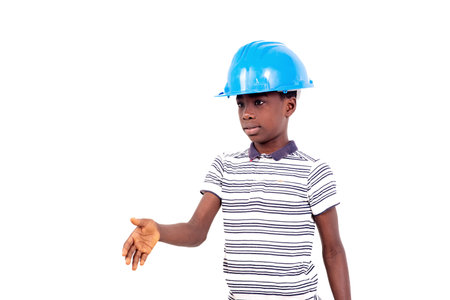 young boy in tee shirt standing on white background wearing safety hat while showing gesture of...