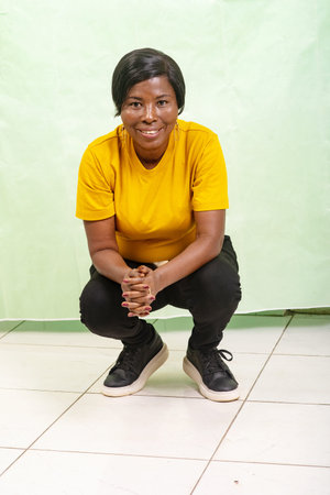 a mature woman in a yellow t-shirt squatting on a green background looking at the camera smiling.