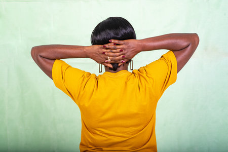 a beautiful mature woman in a yellow t-shirt standing on a green background with her hands on her neck giving her back to the camera.の写真素材