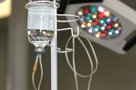 bottle of liquid drug with lines and needle inside called drop counter or drip chamber on a pole in surgery room with lamp in backgroundの写真素材
