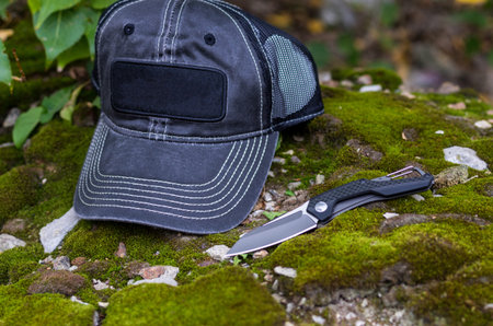 Black folding knife and black baseball cap. Photo on the background of moss.