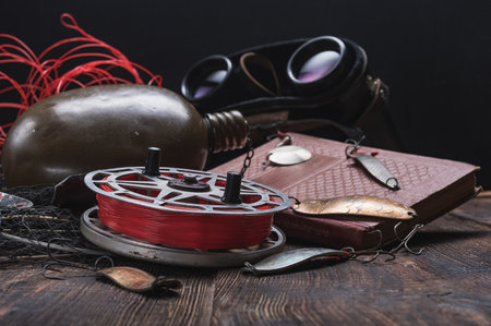 Old vintage fishing reel and baits. Aluminum round fishing reel and red line. Soviet book and binoculars. Low key.の写真素材