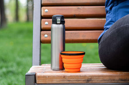 Steel flask and silicone mug. Tourist dishes on the bench. Drinking coffee in the park.の写真素材