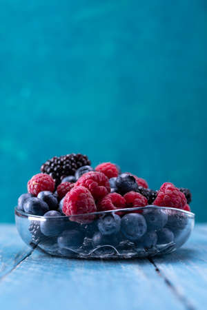 A variety of berries in glassware on a turquoise background. Raspberries, blueberries and blackberries in a bowl. Vertical frame.の写真素材