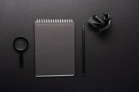Black notepad for notes on a black table. Black pen and magnifier. Black objects on a table. View from above.の写真素材