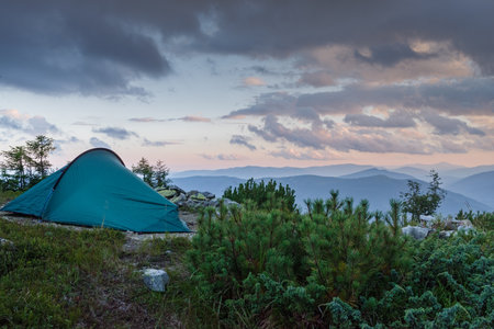 Camping tent on a mountain summit surrounded by rocks and alpine grass. Fiery sunset glows on the horizon with heavy dark clouds above.の写真素材