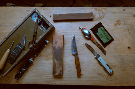 A variety of old rusty knives and hand tools scattered on a weathered wooden table near an open wooden toolbox. Concept of traditional craftsmanship, antique hardware, and workshop mess.の写真素材