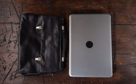 A closed grey laptop lies side by side with a black leather briefcase on a wooden table. Minimalist business workspace setup, remote work concept.