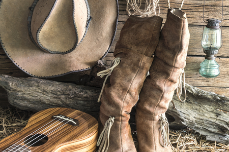 Closeup american west rodeo brown felt cowboy hat and traditional leather boots with ukulele in vintage ranch barn background, Still life styleの写真素材