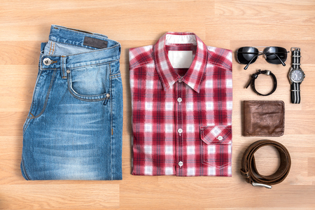 Men's casual outfits with accessories on wooden table, Red plaid shirt and blue jeans with eyeglasses, bracelet, wallet, brown belt and watch, top viewの写真素材