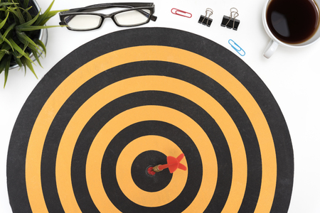 Dart target arrow hitting on bullseye in dartboard over office desk table background with eye glasses, pen, pencil, computer and cup of coffeeの写真素材