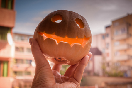 Jack o'Lantern Halloween carved pumpkin smiles widely in orange sunset light with autumnal urban view in the background.の写真素材