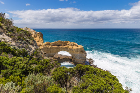 Dramatic cliffs soar over turquoise waters and waves crash beneath the rock formation The Arch along the Great Ocean Road, offering breathtaking views.の写真素材