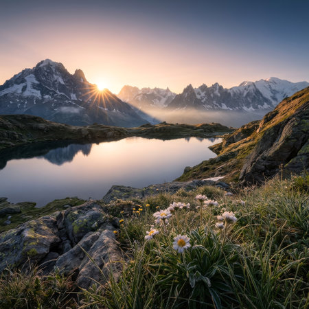 First light illuminates a majestic alpine scene. A radiant sunburst crests jagged peaks, casting a warm glow over a mirror-like lake and blooming wildflowers on the shore.の素材