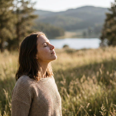 A serene woman with eyes closed breathes deeply, basking in the warm golden hour sun. She stands in a tranquil meadow by a lake, finding a moment of peaceful mindfulness.の素材