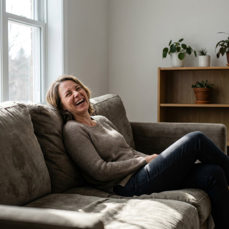 A relaxed woman enjoys a moment of pure happiness, laughing with her eyes closed on a grey sofa. Sunlight streams in, creating a warm, cozy atmosphere.の素材