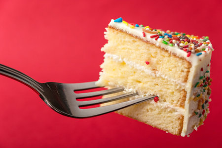A slice of vanilla cake with white frosting and sprinkles on a fork, against redの素材
