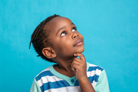 Young boy in overalls thoughtfully gazes upward against a blue backdrop.の素材