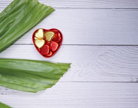 valentine day concept. red and gold mini heart in heart shape box with green leaves. white wood texture backgroundの写真素材