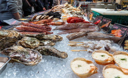 A market scene shows a large display of fresh seafood items on ice. Shellfish, fish, and other seafood varieties are arranged for customers. People are browsing nearby.の写真素材