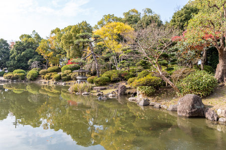 People stroll on the pathway beside a calm pond surrounded by trees in a Japanese garden. The season is autumn with colorful leaves and clear skies.の写真素材