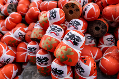 Daruma dolls are stacked together at a market in Japan during a festival. Each doll shows unique facial features and colors. People walk by admiring the art.の写真素材