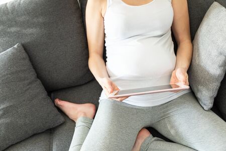Young pregnant woman holds her belly and looks at her tablet sitting on a sofa
