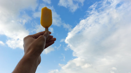 Male hand holding yellow ice cream stick against blue sky with white cloudsの写真素材
