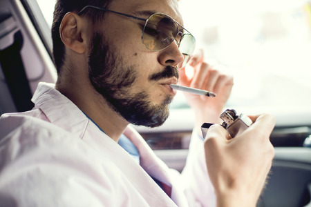 Young businessman lighting up a cigarette with a lighter in his cupped hands puffing smoke.の写真素材