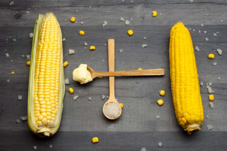 Two cobs of fresh and cooked sweet corn lying on a gray table surrounded by salt and butter on a wooden spoon.の写真素材